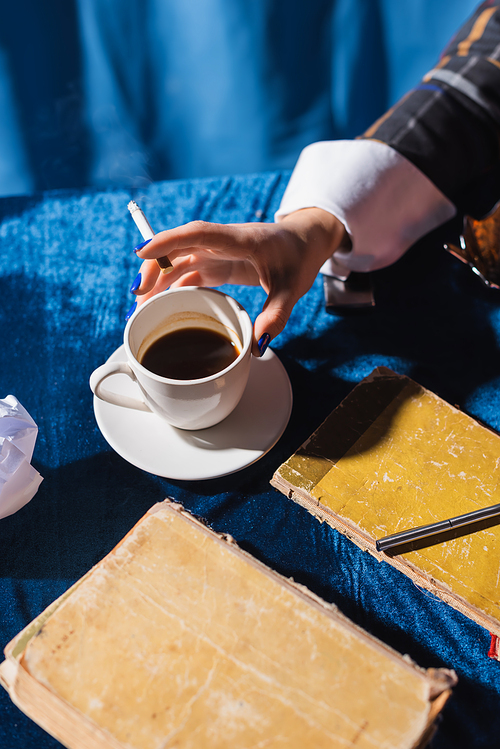 partial view of woman with cigarette near cup of coffee and vintage books on blue velour tablecloth