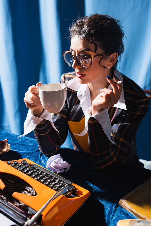 vintage style newswoman in eyeglasses smoking and drinking coffee near typewriter on blue background