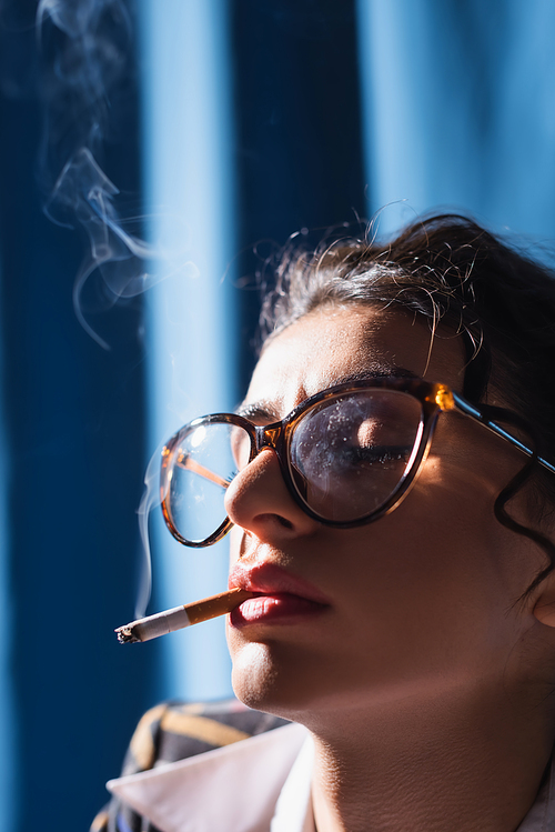 close up view of woman in vintage eyeglasses smoking with closed eyes on blue background