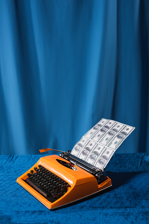red vintage typewriter with dollars on velour table near blue drape on background