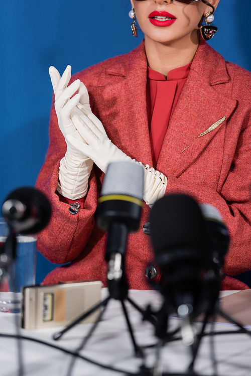 partial view of stylish woman in red blazer and white gloves near blurred microphones on blue background