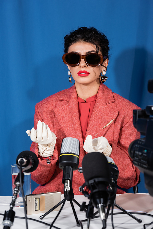 young woman in vintage sunglasses gesturing during interview on blue background