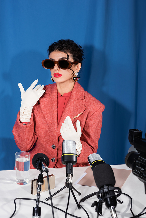 stylish woman in vintage sunglasses and white gloves gesturing during interview on blue background