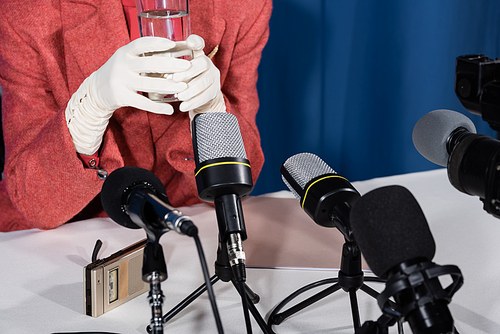 cropped view of woman in white gloves holding glass of water near microphones and dictaphone on blue background