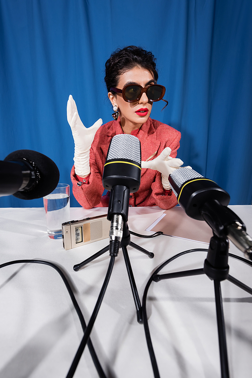 microphones near vintage style woman in sunglasses gesturing during interview on blue background