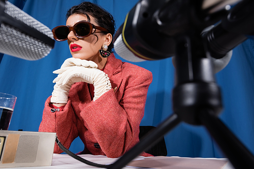 blurred microphones near stylish woman in vintage sunglasses and white gloves on blue background