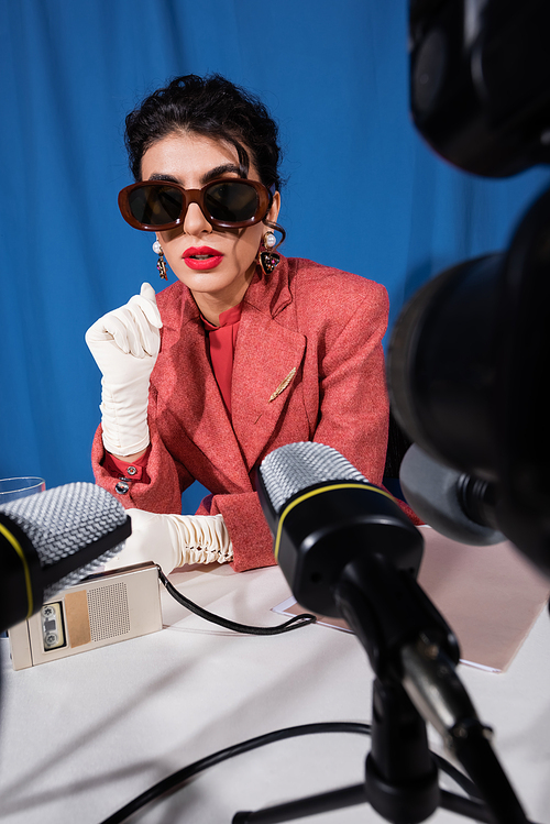 retro style woman in sunglasses talking near blurred microphones on blue background
