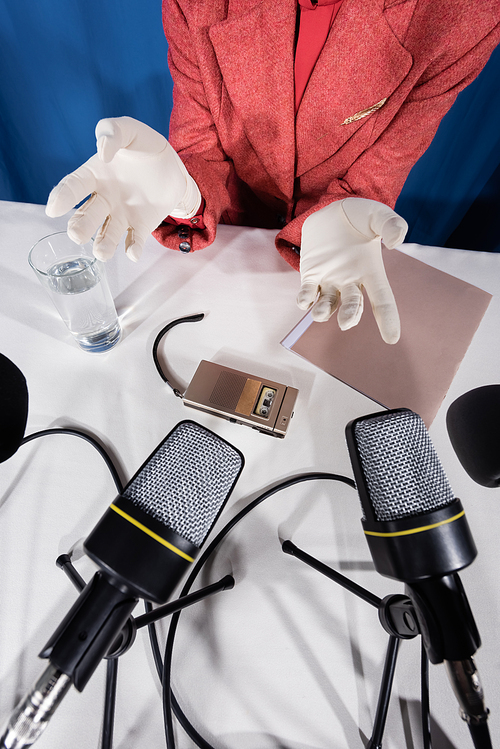 top view of microphones, dictaphone and glass of water near cropped woman in white gloves