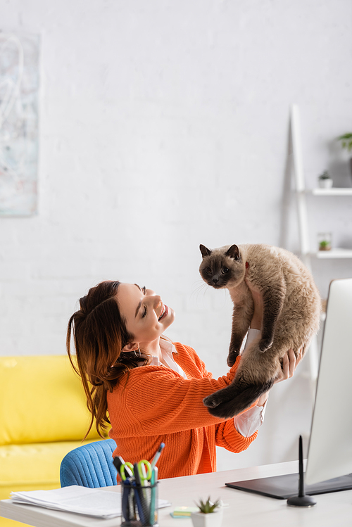 happy woman holding cat while sitting at work desk at home