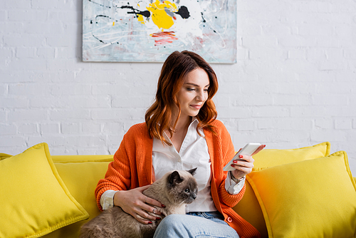 smiling woman messaging on mobile phone while sitting on yellow couch with cat