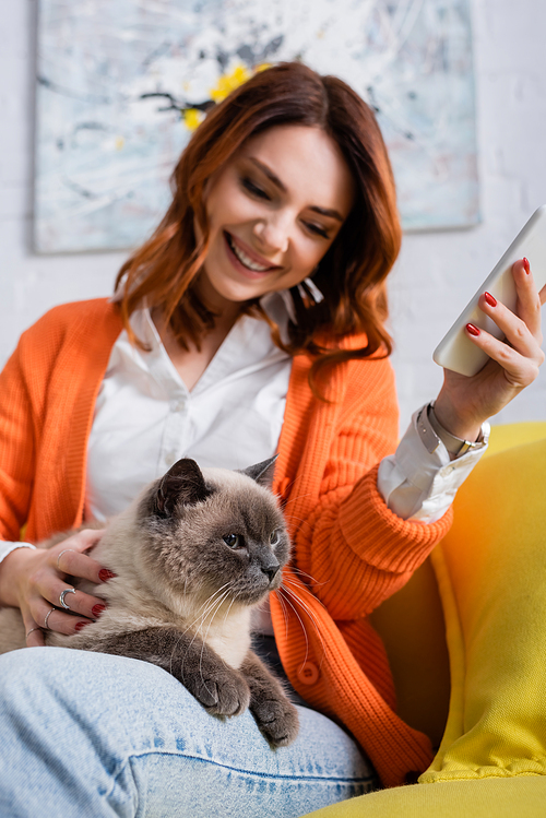 happy woman cuddling cat while sitting on couch with mobile phone, burred background