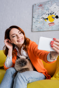 happy woman fixing hair while sitting on couch and taking selfie with cat