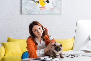 cheerful woman in headset  while working near cat on desk at home