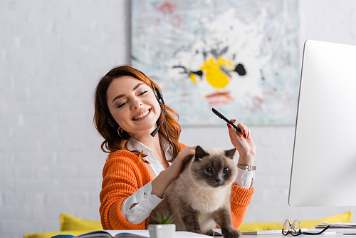 cheerful woman in headset stroking cat while working near blurred computer monitor