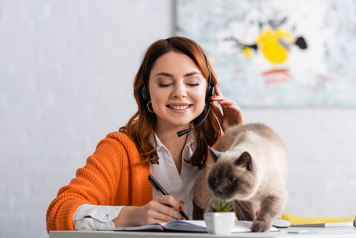 happy woman in headset writing in notebook near blurred cat on work desk