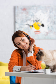 cheerful woman in headset writing in notebook while working near cat sitting on desk