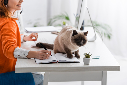 cropped view of blurred woman writing in notebook near cat sitting on work desk