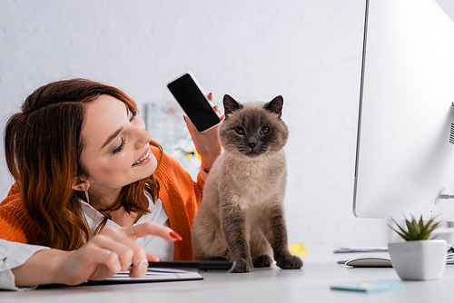 smiling freelancer holding smartphone with blank screen near cat sitting on desk near computer monitor