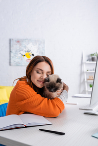 happy woman embracing cat while sitting near work desk at home