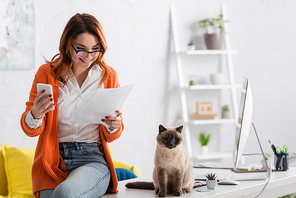 cheerful freelancer with smartphone looking at documents near cat sitting on desk