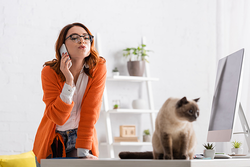 woman in eyeglasses pouting lips while calling on cellphone near blurred cat on desk