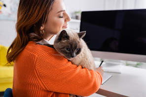 pleased woman with closed eyes hugging cat near blurred monitor with blank screen