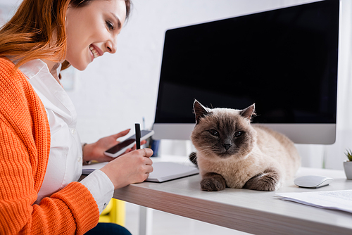 smiling woman with smartphone writing in notebook near cat on desk