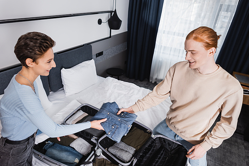 High angle view of smiling couple holding clothes near suitcases on bed in hotel room