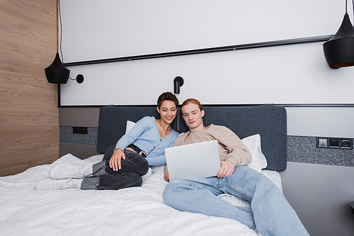 Cheerful couple using laptop on bed in hotel room