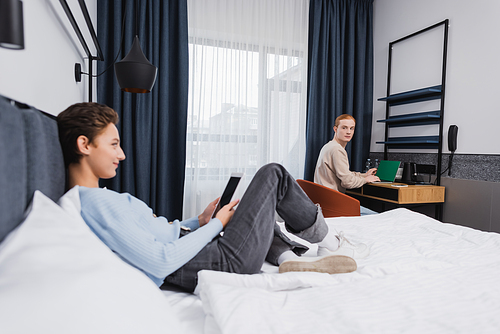 Young man using laptop near blurred girlfriend with digital tablet in hotel room