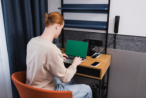 Redhead man using laptop with green screen near smartphone in hotel room