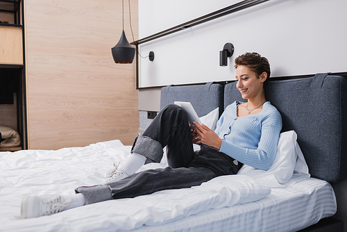 Positive woman using digital tablet on bed in hotel room