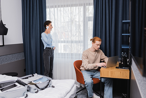 Smiling woman talking on smartphone near boyfriend using laptop and suitcase on bed in hotel