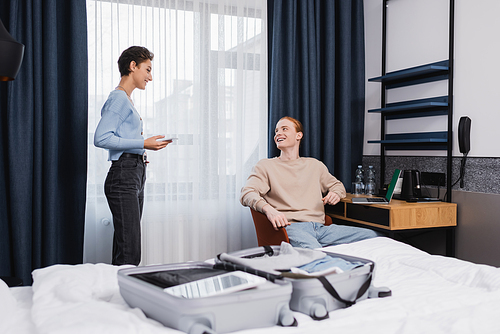 Positive young couple with gadgets talking near suitcase on bed in hotel room