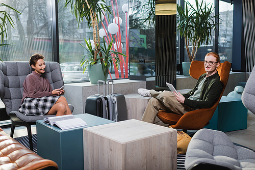 Positive couple using devices near suitcases in hotel lobby