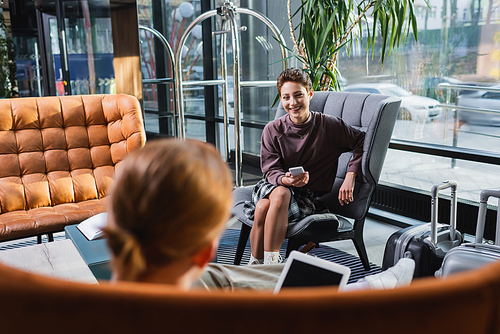 Smiling woman holding smartphone near blurred boyfriend and suitcases in hotel lobby