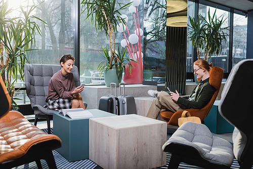 Smiling woman using smartphone near boyfriend with digital tablet and suitcases in hotel lobby