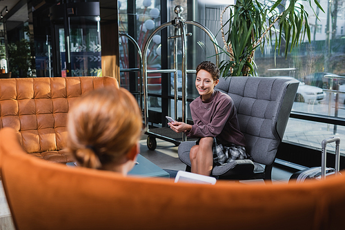 Smiling woman with cellphone looking at blurred boyfriend in hotel lobby