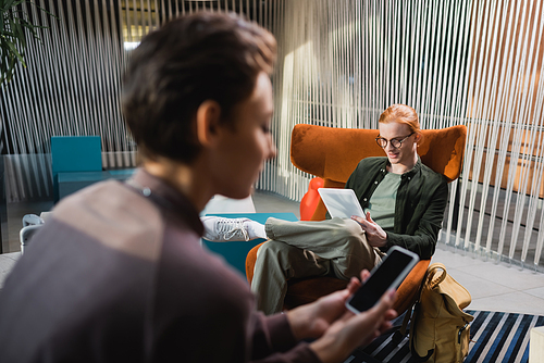 Young man using digital tablet near blurred girlfriend with smartphone in hotel lobby