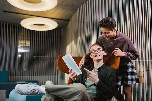 Smiling man pointing at digital tablet near girlfriend in hotel lobby