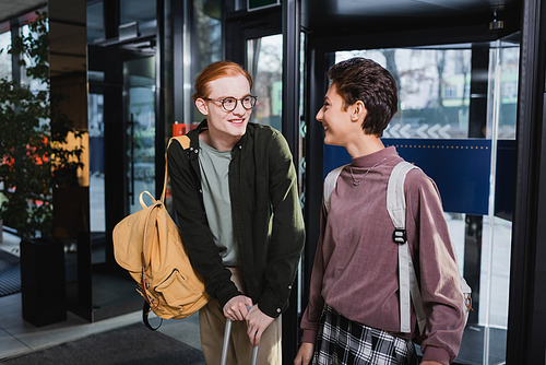 Cheerful man with suitcase and backpack looking at girlfriend in hotel