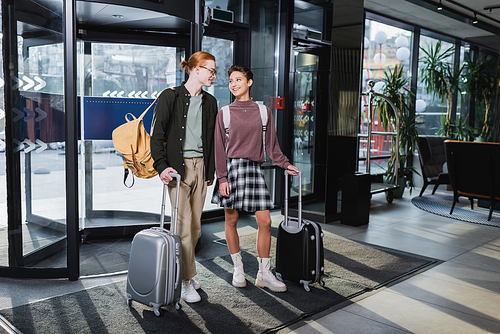 Smiling woman with backpack and suitcase standing near boyfriend in hotel lobby