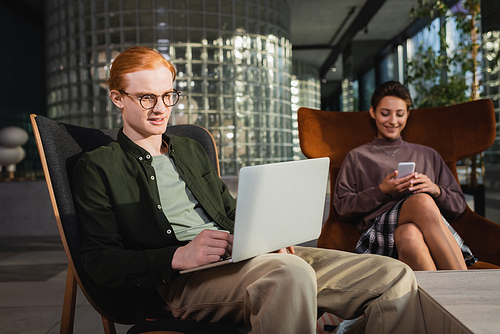 Smiling freelancer using laptop near blurred girlfriend with smartphone in hotel lobby