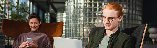 Young man looking at laptop near blurred girlfriend with smartphone in hotel lobby, banner