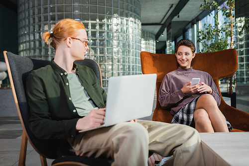 Smiling woman with cellphone looking at blurred boyfriend using laptop in hotel lobby