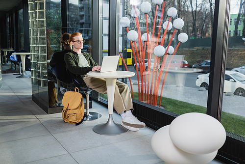 Redhead freelancer using laptop near backpack in hotel lobby