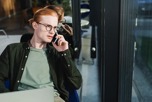 Redhead man talking on smartphone near blurred laptop in hotel lobby