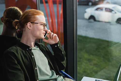 Side view of young freelancer talking on smartphone near laptop in hotel