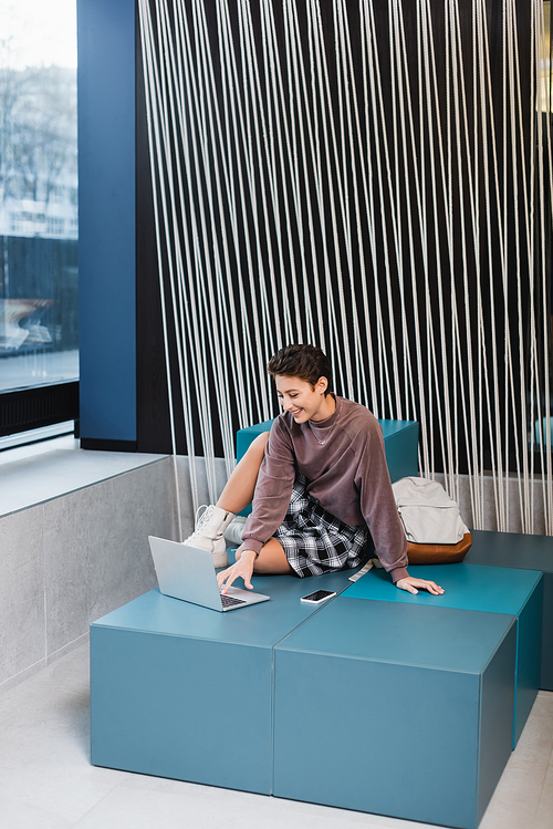 Smiling traveler using laptop near backpack in modern hotel lobby