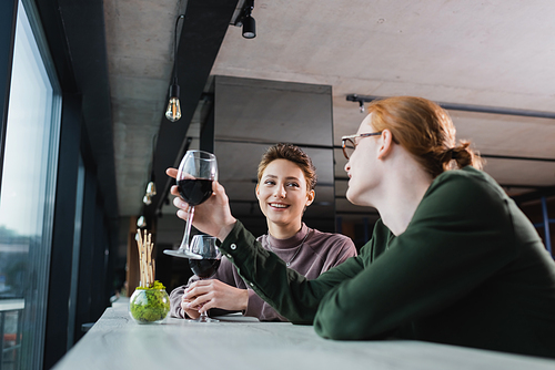 Cheerful woman looking at blurred boyfriend with wine in hotel